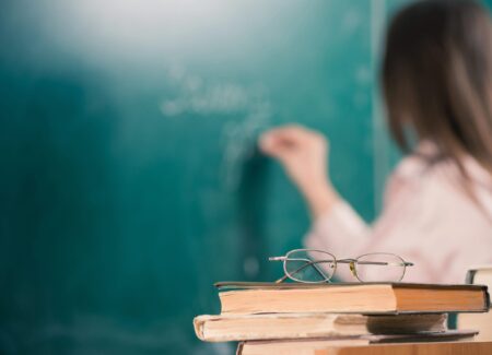 Teacher writing on a green chalkboard with glasses on top of a stack of books in the foreground.